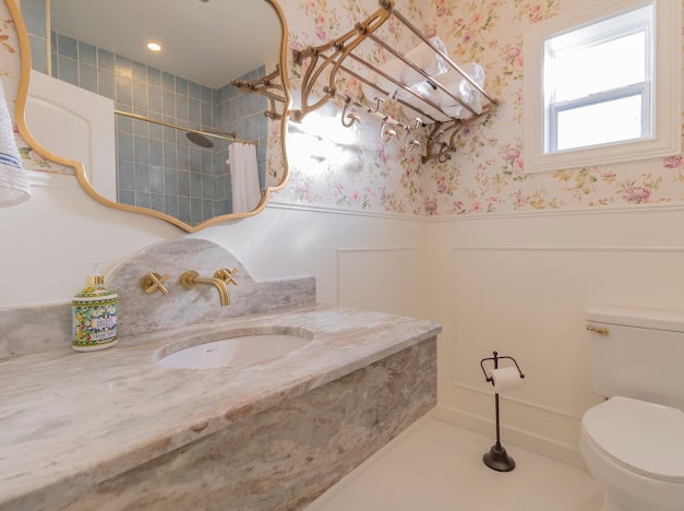 A well-appointed bathroom features floral wallpaper that adds a touch of charm. A marble countertop surrounds a round sink, while a large mirror reflects the space. Fixtures in a polished finish complement the elegant design, and a window provides natural light.