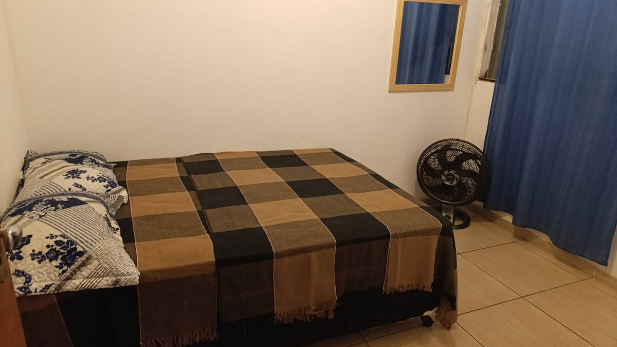 A private bedroom is featured, furnished with a spacious bed covered by a checker-patterned blanket. Two pillows are placed at the head of the bed. A small fan and a mirror are also present, while light blue curtains hang beside a window.