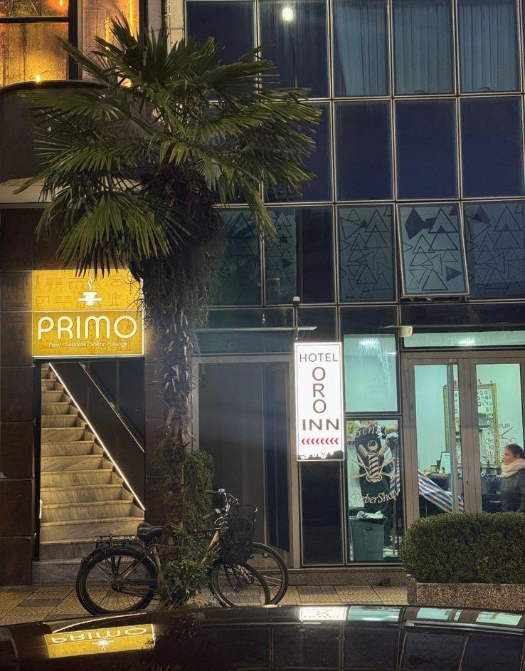 A building facade features the illuminated sign of 'Hotel Oro Inn' beside a palm tree. The entrance includes glass doors, offering visibility into the space. A bicycle is parked nearby, emphasizing a blend of urban life and casual accessibility.