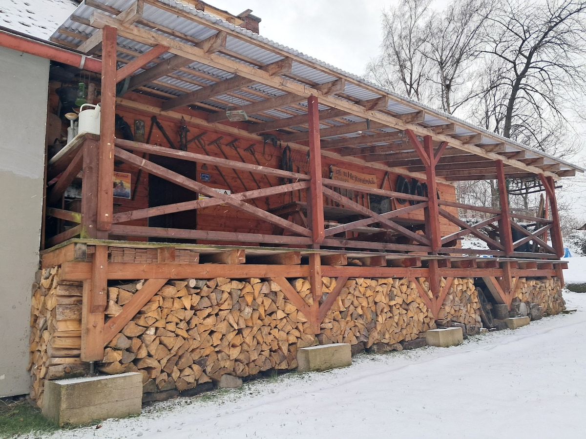 A rustic wooden terrace is supported by sturdy posts, featuring a safety railing and ample space for seating. Stacked firewood is neatly arranged beneath the raised platform, while a snow-covered landscape surrounds the space, enhancing the serene atmosphere.