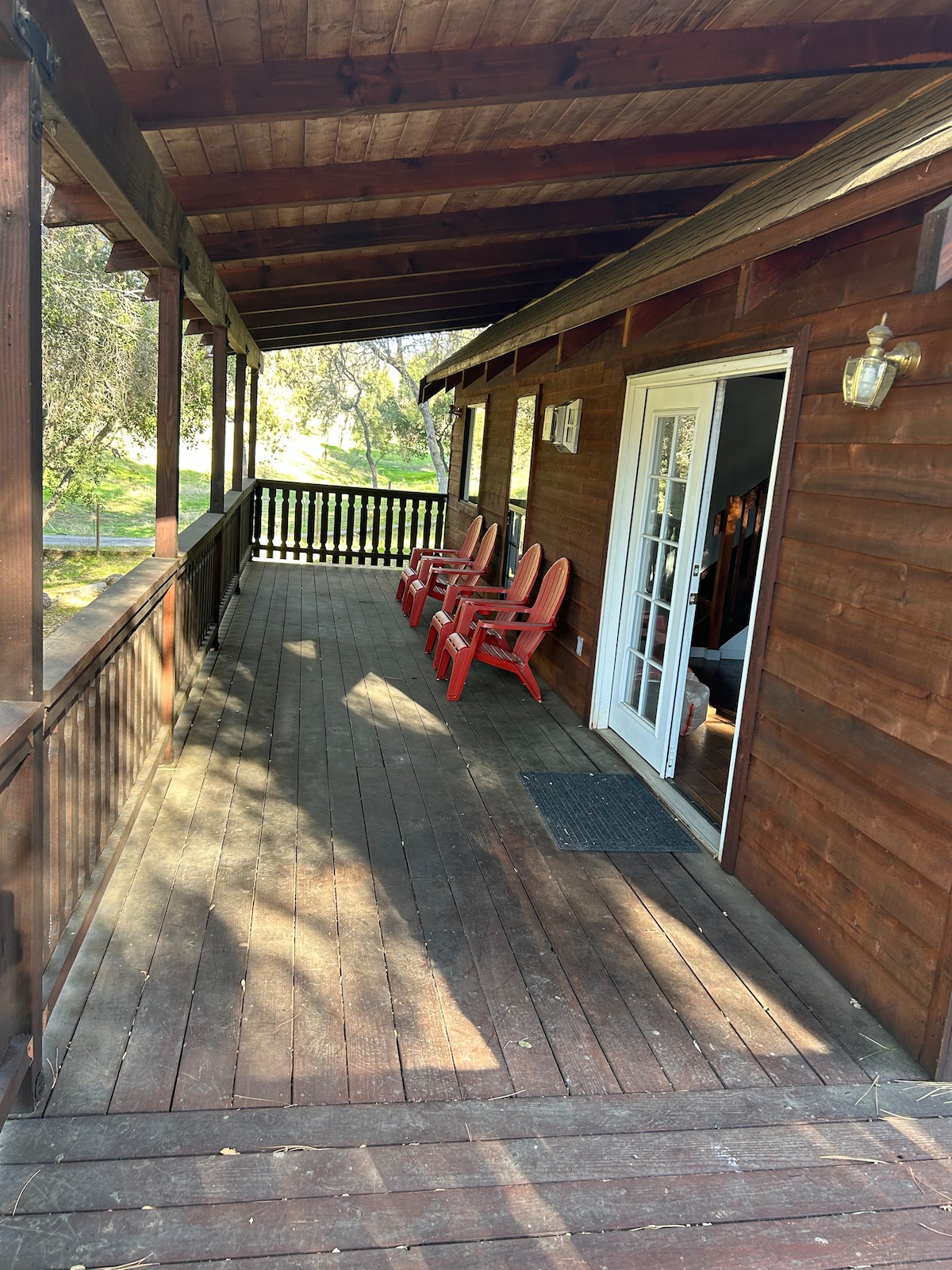 A spacious cabin porch features wooden flooring and a rustic wooden railing. Several red Adirondack chairs are positioned along the edge, offering a relaxing spot. Sunlight streams onto the porch, enhancing the warm ambiance of the setting.