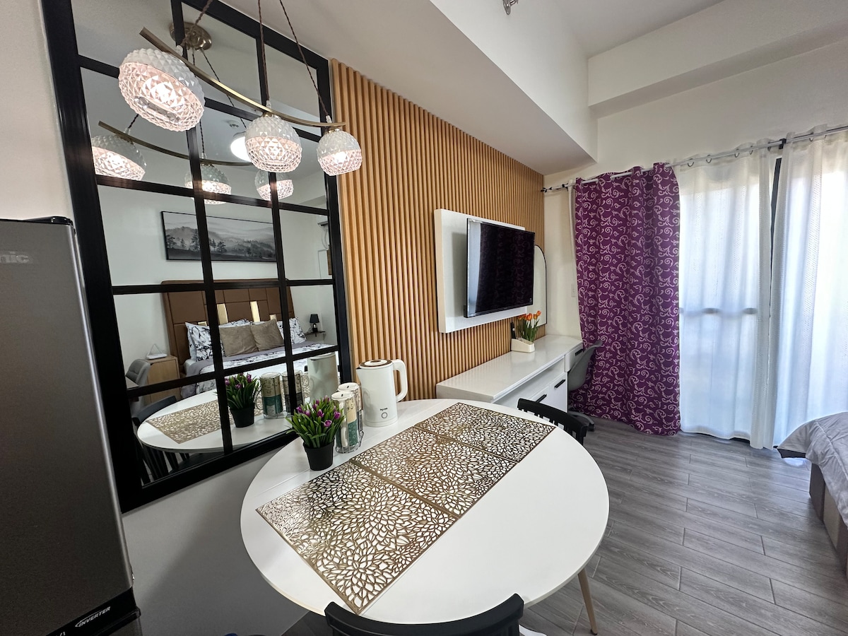 A dining area features a round table with elegant placemats and modern chairs. A mirror behind the table reflects light fixtures, enhancing the sense of space. A television is mounted on a wall, and light filters through sheer curtains, complemented by patterned drapes.