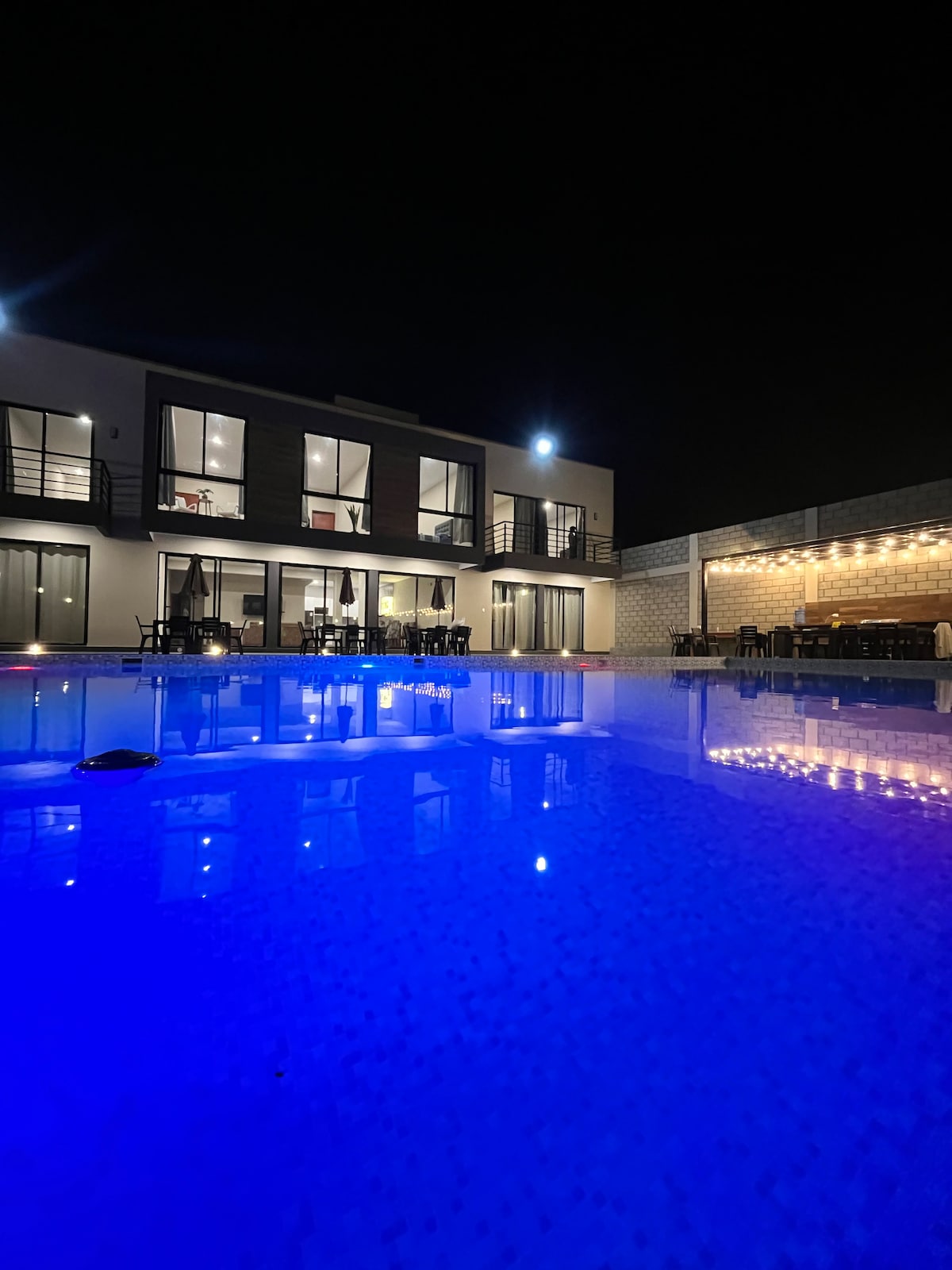 A modern house is illuminated at night, reflecting on a sparkling blue pool. The exterior features large windows, providing a view of the interior activities. A patio area with string lights creates a welcoming ambiance for outdoor gatherings.
