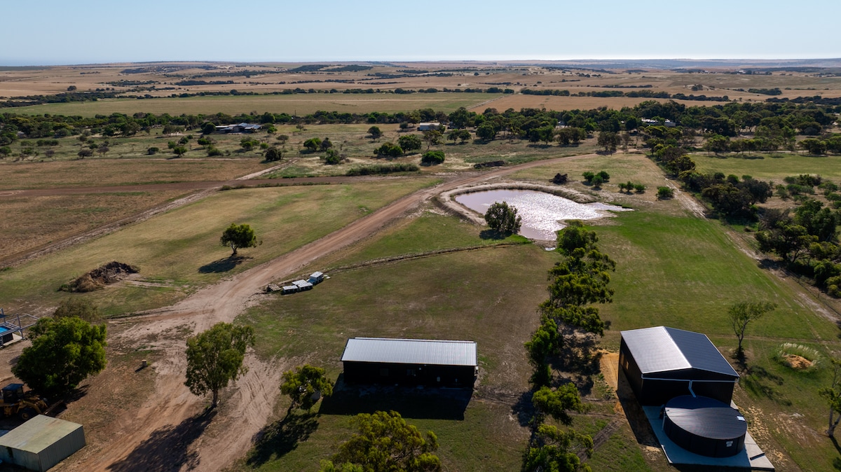 Aerial view of a spacious countryside landscape featuring open fields and patches of trees. A small pond reflects the surroundings, while distant coastal views are visible. Several structures, including sheds, are scattered throughout the area, contributing to a serene rural setting.
