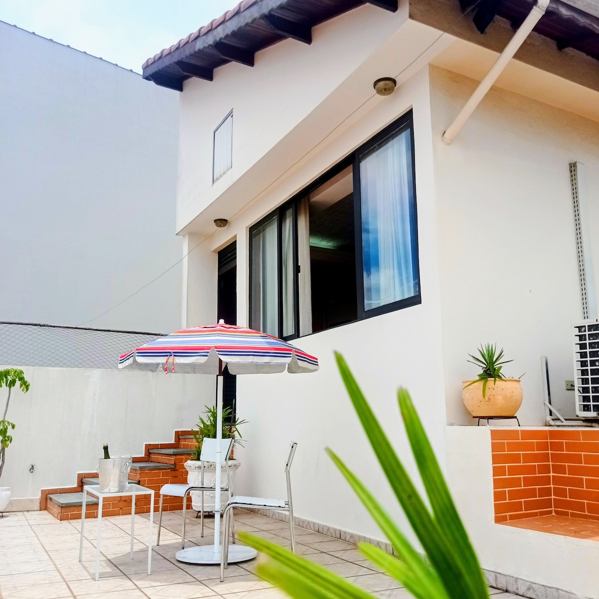 An outdoor terrace is showcased with a vibrant striped umbrella and a small table and chair set. The airy exterior of the guest house can be seen, featuring large windows that brighten the interior space. Potted plants add a touch of greenery.