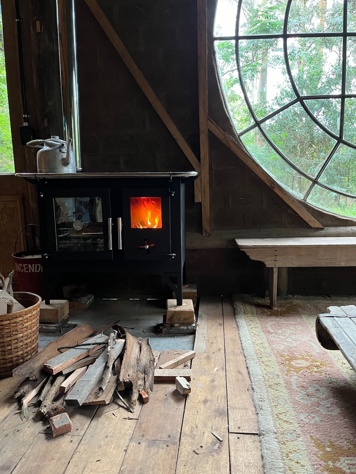 A wood-burning stove stands prominently in the corner, with warm flames visible through the glass door. Nearby, a stack of firewood is neatly arranged on the wooden floor. A large round window provides a view of the surrounding greenery, enhancing the rustic charm of the room.