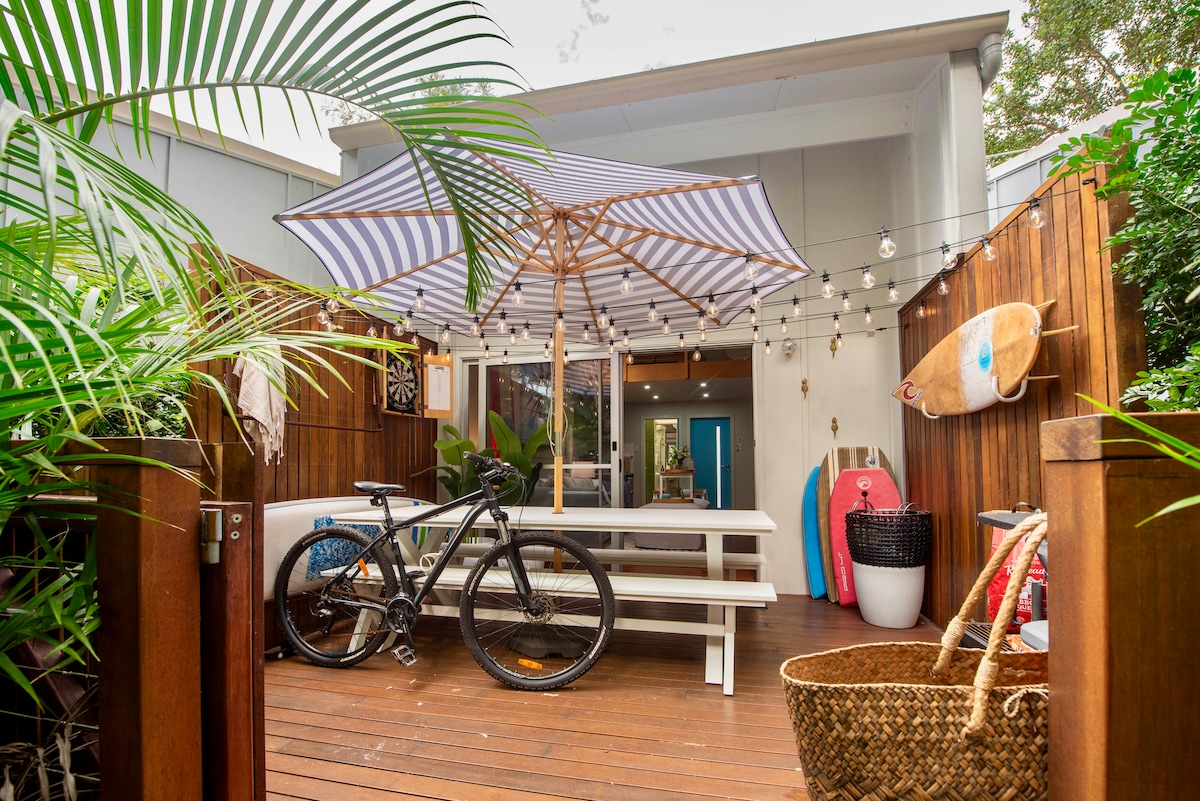An outdoor deck area is showcased, featuring a large striped umbrella over a dining table. A mountain bike rests nearby, alongside colorful surfboards. Lush greenery and decor items such as a large basket and wall art enhance the inviting space.