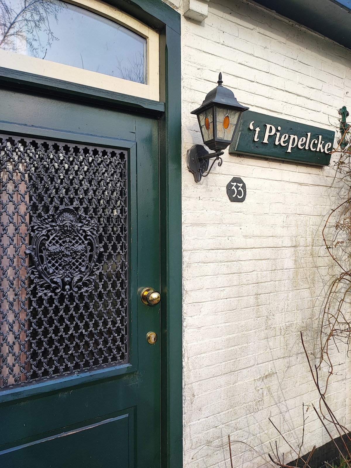 The entrance to 't Piepelcke features a green door with an intricate wrought iron design. A lantern is mounted beside the door, illuminating the white brick wall. The building number 33 is displayed, indicating the main access point to the charming property.