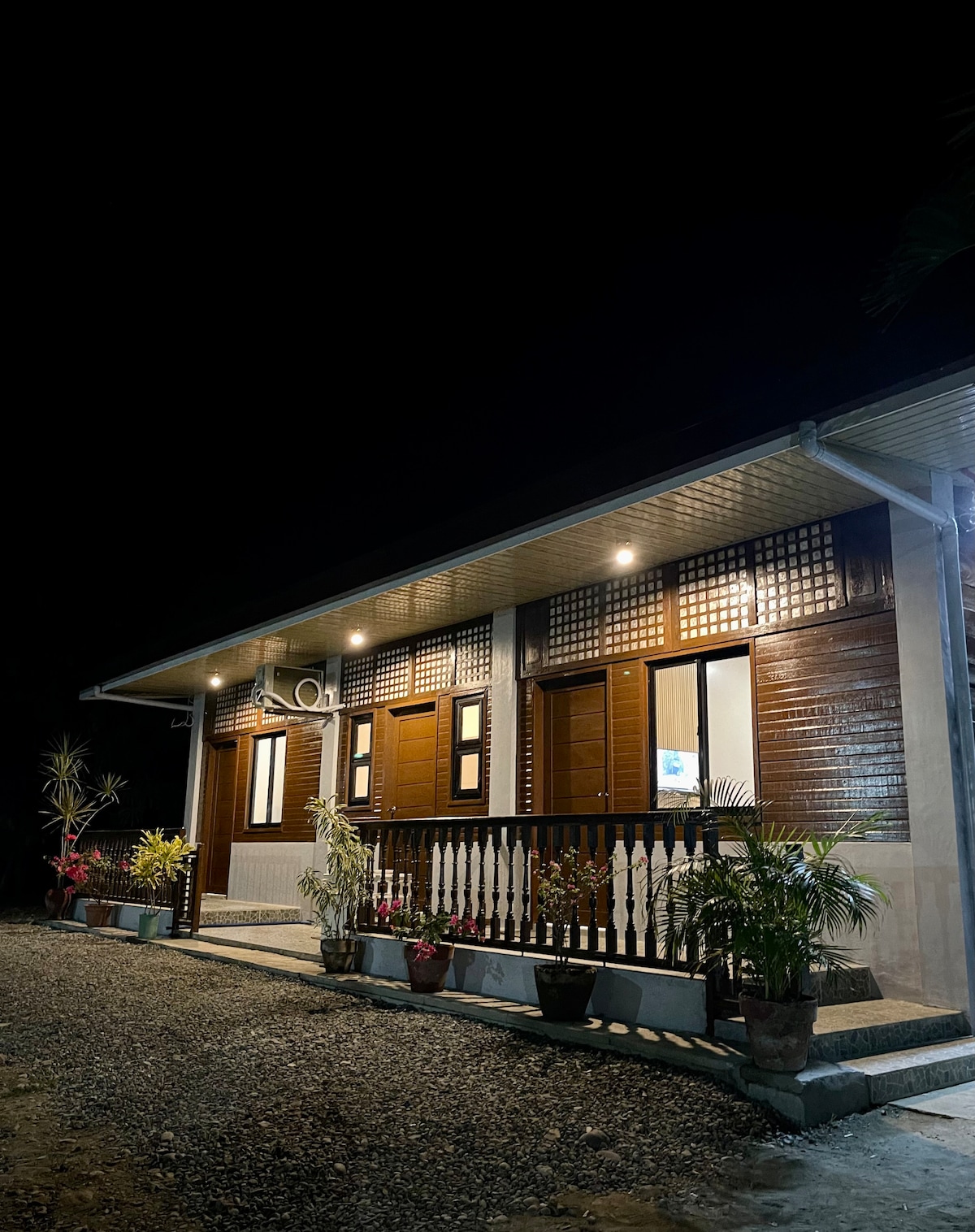 The exterior of the cabin is illuminated by soft outdoor lighting, highlighting the wooden architecture and decorative balcony. Potted plants add a touch of greenery along the entrance, while large windows allow glimpses of the interior. A gravel path leads up to the welcoming entry.