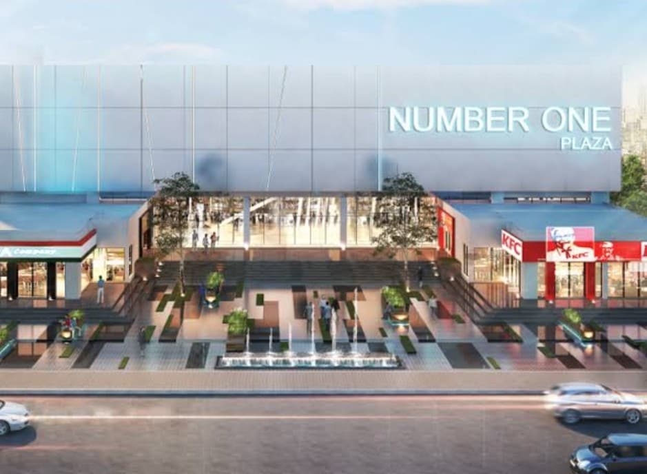 A modern shopping plaza is visible, featuring a sleek glass facade and prominent signage reading 'NUMBER ONE PLAZA.' Steps lead to a spacious entrance adorned with potted plants and water features. Fast-food outlets are situated on either side, enhancing the bustling environment.