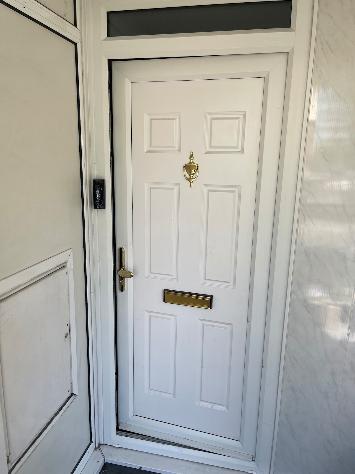A white front door is displayed, featuring a brass knocker and a mail slot. The door is set within a marble-patterned wall. Beside the door, a small intercom or keypad is visible, enhancing the entryway's accessibility.