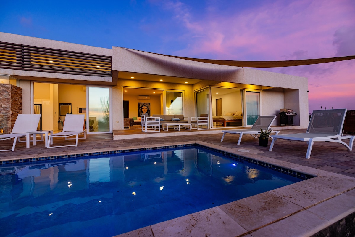 A modern villa features a private pool surrounded by light lounge chairs. Large glass doors open to a spacious living area illuminated by warm lighting. The evening sky showcases purple and orange hues, contributing to a tranquil ambiance.