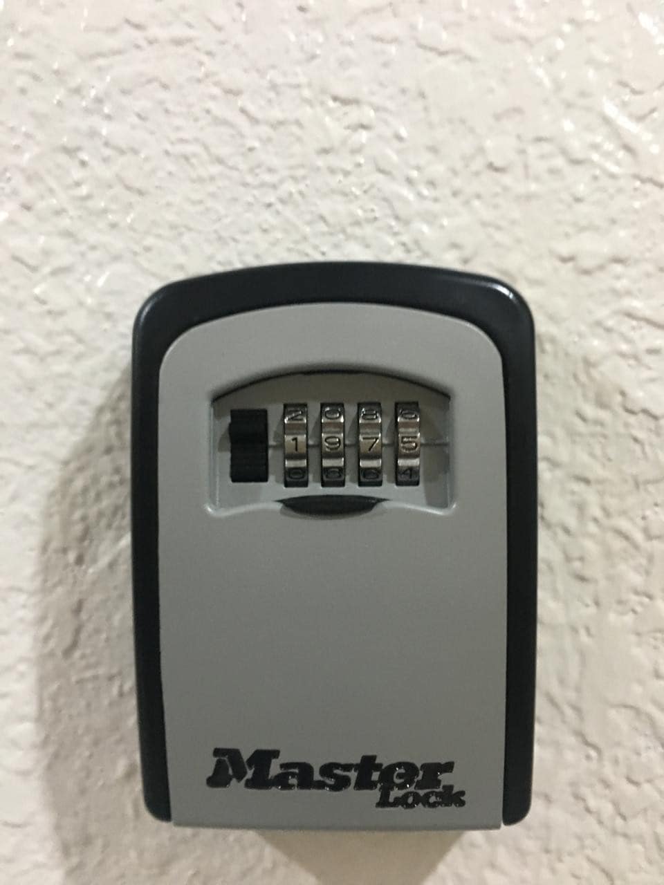 A wall-mounted lockbox is displayed, featuring a combination dial with four adjustable dials for securing items. The box has a sleek gray and black design, ensuring easy visibility against the textured white wall.
