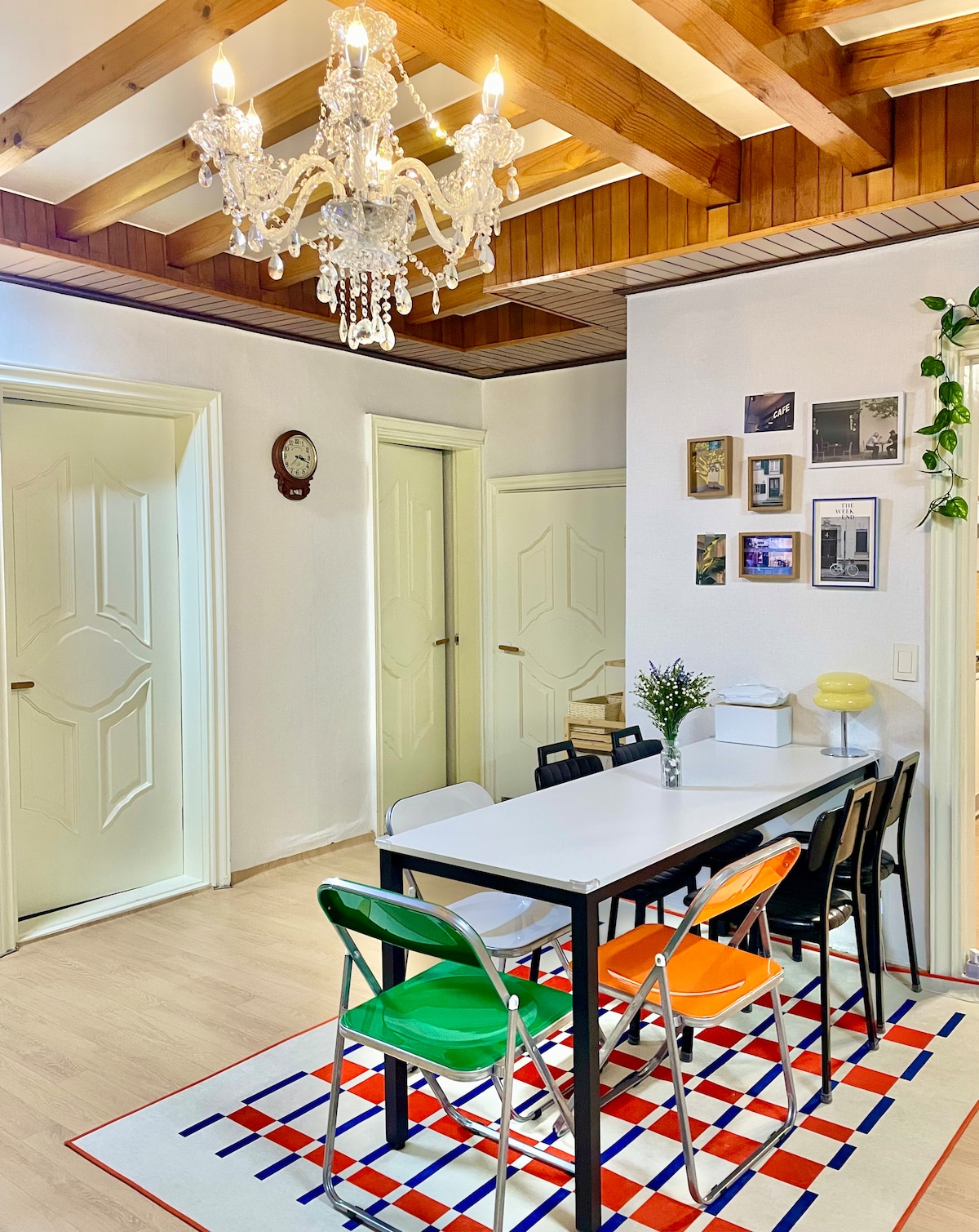 A dining area features a black table surrounded by colorful chairs in green, orange, and white. A crystal chandelier hangs from the wooden ceiling, while framed photographs and a clock adorn the walls. A patterned rug adds a touch of warmth to the space.