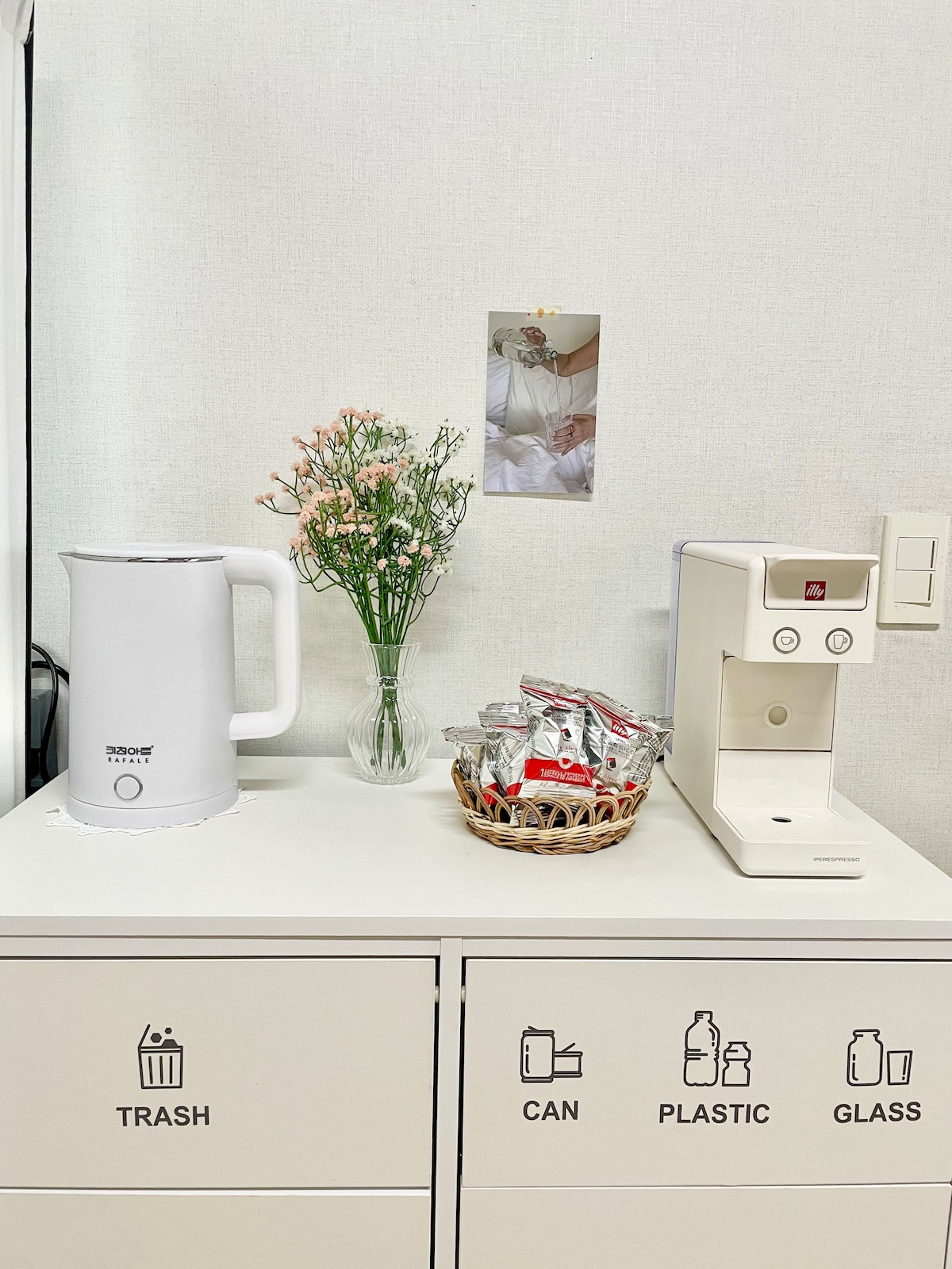 A countertop space features a white electric kettle and a coffee machine positioned beside a small woven basket containing snacks. A clear glass vase holds fresh flowers, adding a touch of greenery. Below, organized bins are labeled for trash, cans, plastic, and glass.