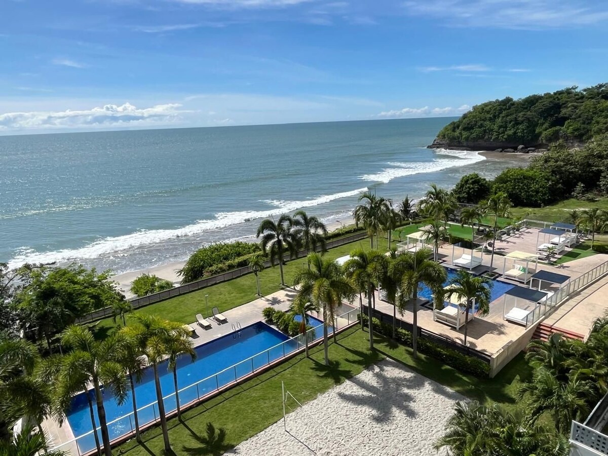 An oceanfront view presents gentle waves lapping against the shore. A spacious pool area is visible, surrounded by lush palm trees and lounging areas. Sunbeds are positioned beside the pool, while a sandy area is noted nearby, enhancing the serene coastal environment.