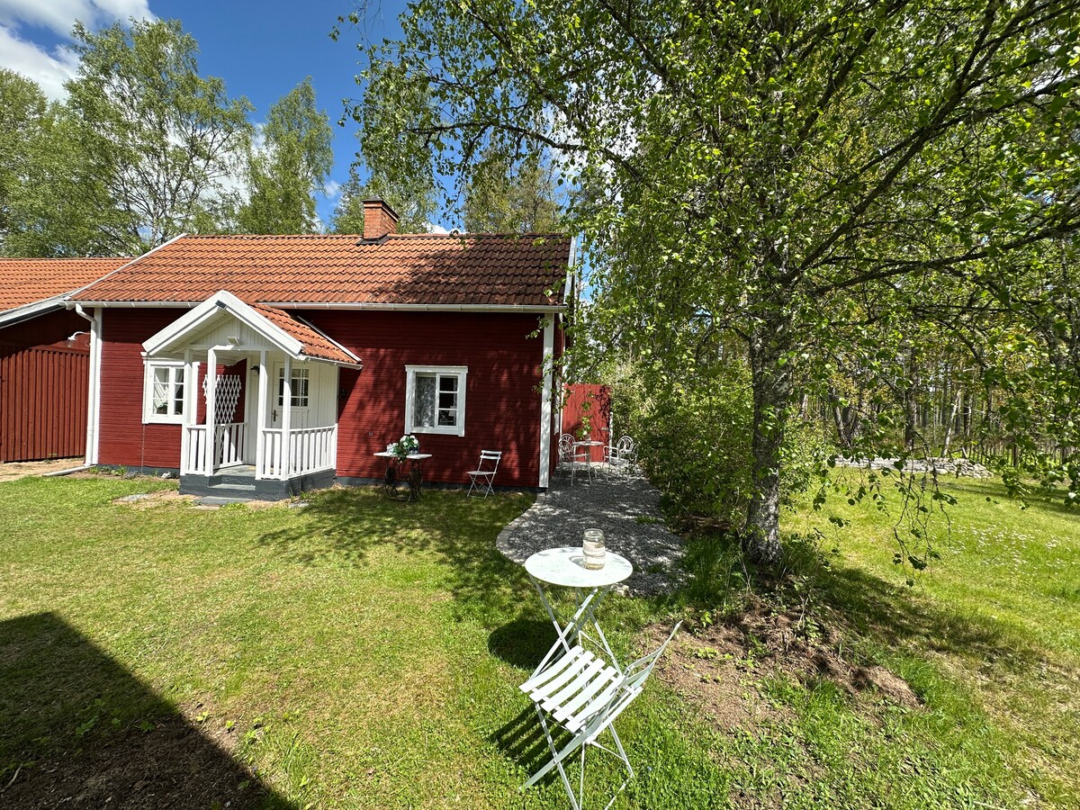 A traditional red Swedish house is surrounded by green grass and trees. A small table and chair are set up on a gravel area, ideal for enjoying the outdoors. The cozy entrance features a white porch, enhancing the welcoming appearance of the home.