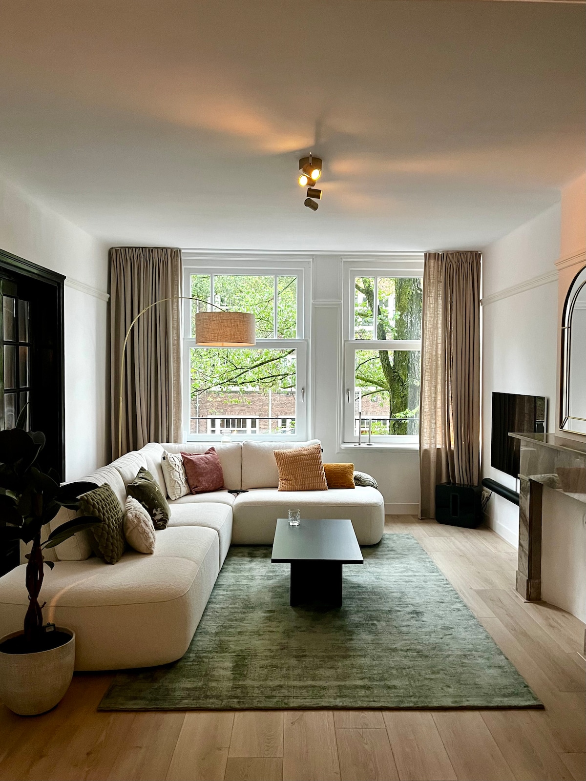 A bright living room is showcased, featuring a spacious white sectional sofa adorned with cushions in warm tones. A sleek black coffee table is positioned on a soft green area rug. Large windows invite natural light, framed by elegant drapes, while greenery is visible outside.