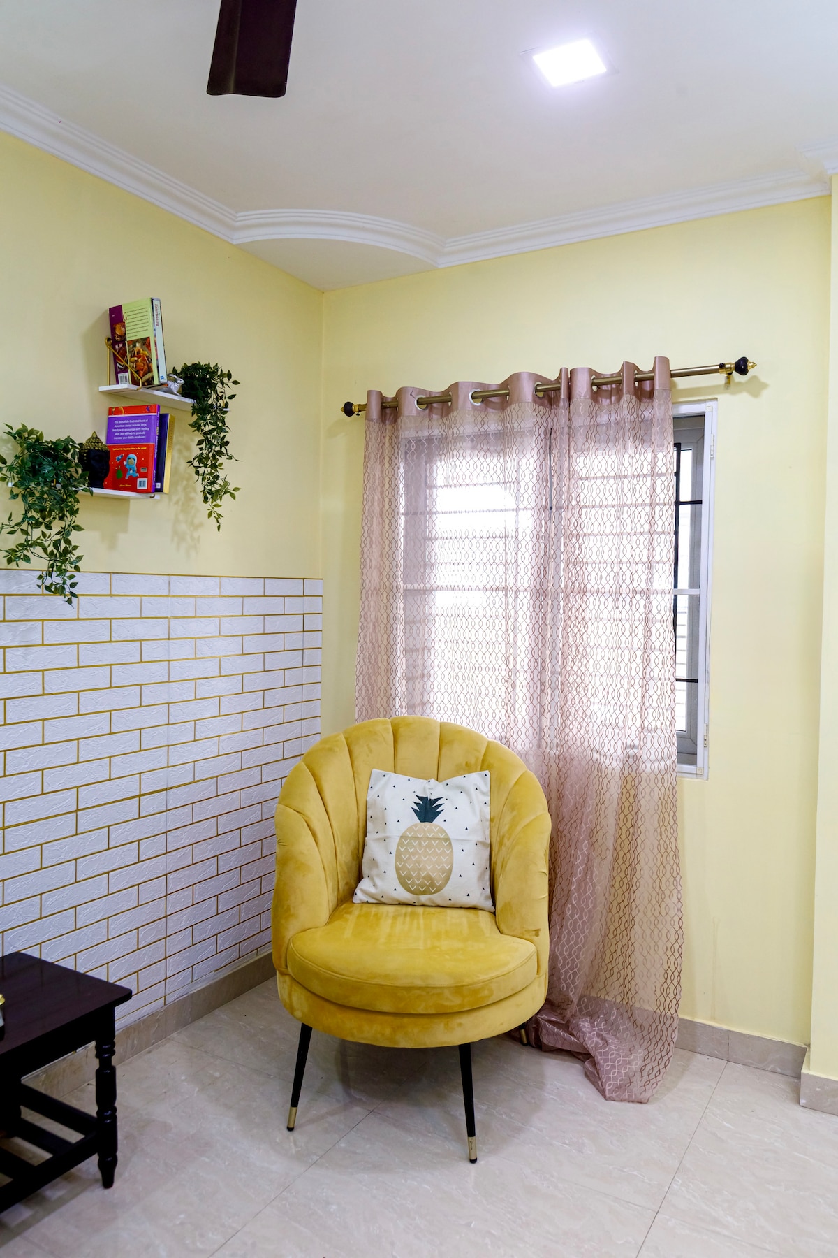 A cozy reading corner features a yellow upholstered chair with a pineapple-patterned cushion. Light streams through a sheer curtain, enhancing the warm yellow walls. A small shelf displays potted plants and decorative books, adding charm to the space.