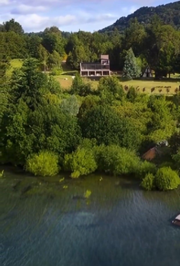 The image captures a serene lakeside view featuring abundant greenery and a backdrop of rolling hills. A charming, multi-level building is visible, nestled among the trees, with a well-maintained lawn leading to the water's edge.