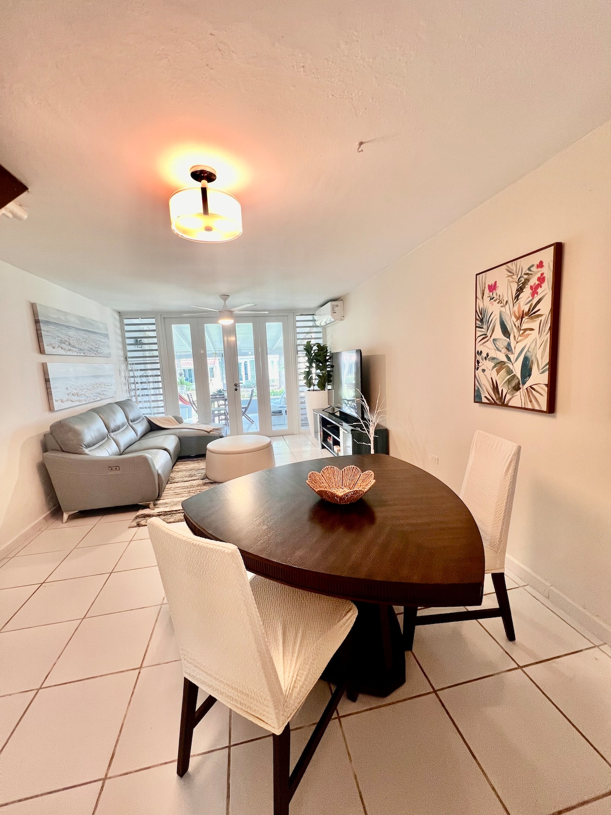 A modern living space is highlighted with a comfortable gray sofa and a circular coffee table. A stylish dining table with two woven chairs is positioned beside the open area. Natural light flows through multiple glass doors, enhancing the fresh, bright atmosphere.