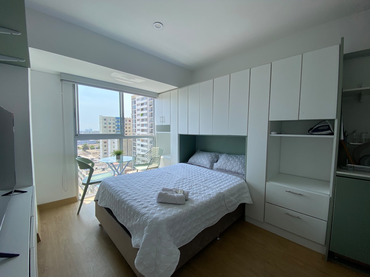 A comfortable bedroom features a spacious bed dressed in white linens, complemented by built-in cabinetry and a cozy reading nook. Large windows offer natural light and a view of the surroundings, while a small table with two chairs is positioned near the window.