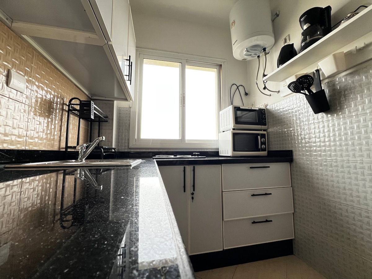 A modern kitchen is showcased with sleek black countertops and white cabinetry. Essential appliances, including a microwave and a kettle, are visible. Natural light enters through a window, enhancing the functional design, which features open shelving and ample workspace.