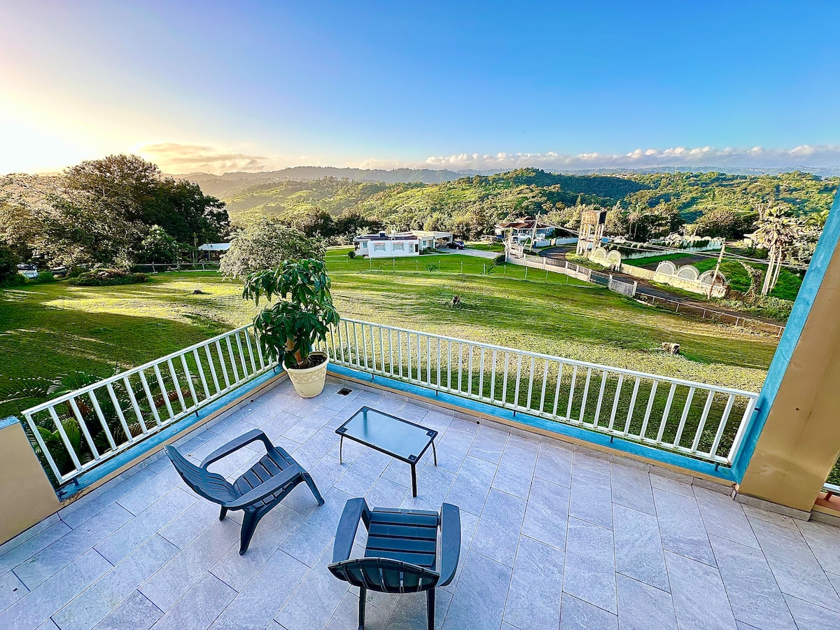 A balcony area features two black chairs positioned around a glass table, providing a comfortable space to enjoy the scenic view. Lush greenery and rolling hills extend in the background, with a clear blue sky above, creating a peaceful atmosphere.
