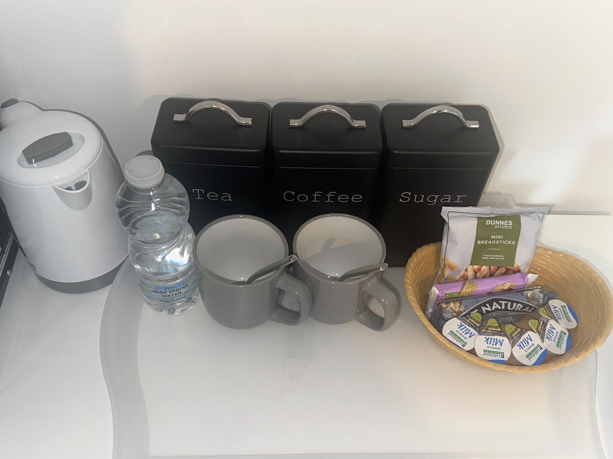A tea and coffee station is arranged on a surface, featuring a kettle, water bottle, and three canisters labeled for tea, coffee, and sugar. Two grey mugs sit alongside a woven basket containing creamers and assorted snacks.