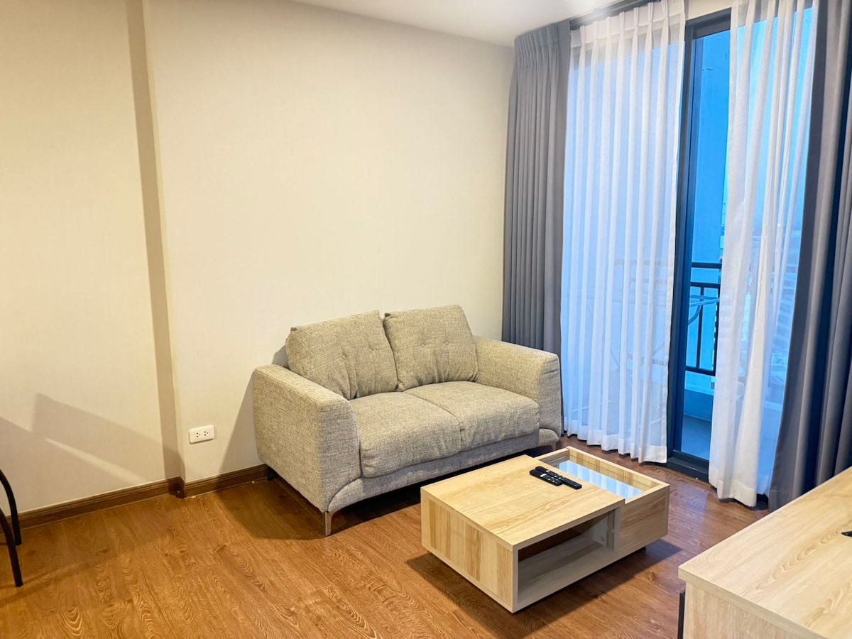 A comfortable seating area features a light gray sofa positioned against a neutral wall. A wooden coffee table sits in front, with a remote control on its surface. Sheer white curtains frame a balcony door, allowing natural light to enter the space.