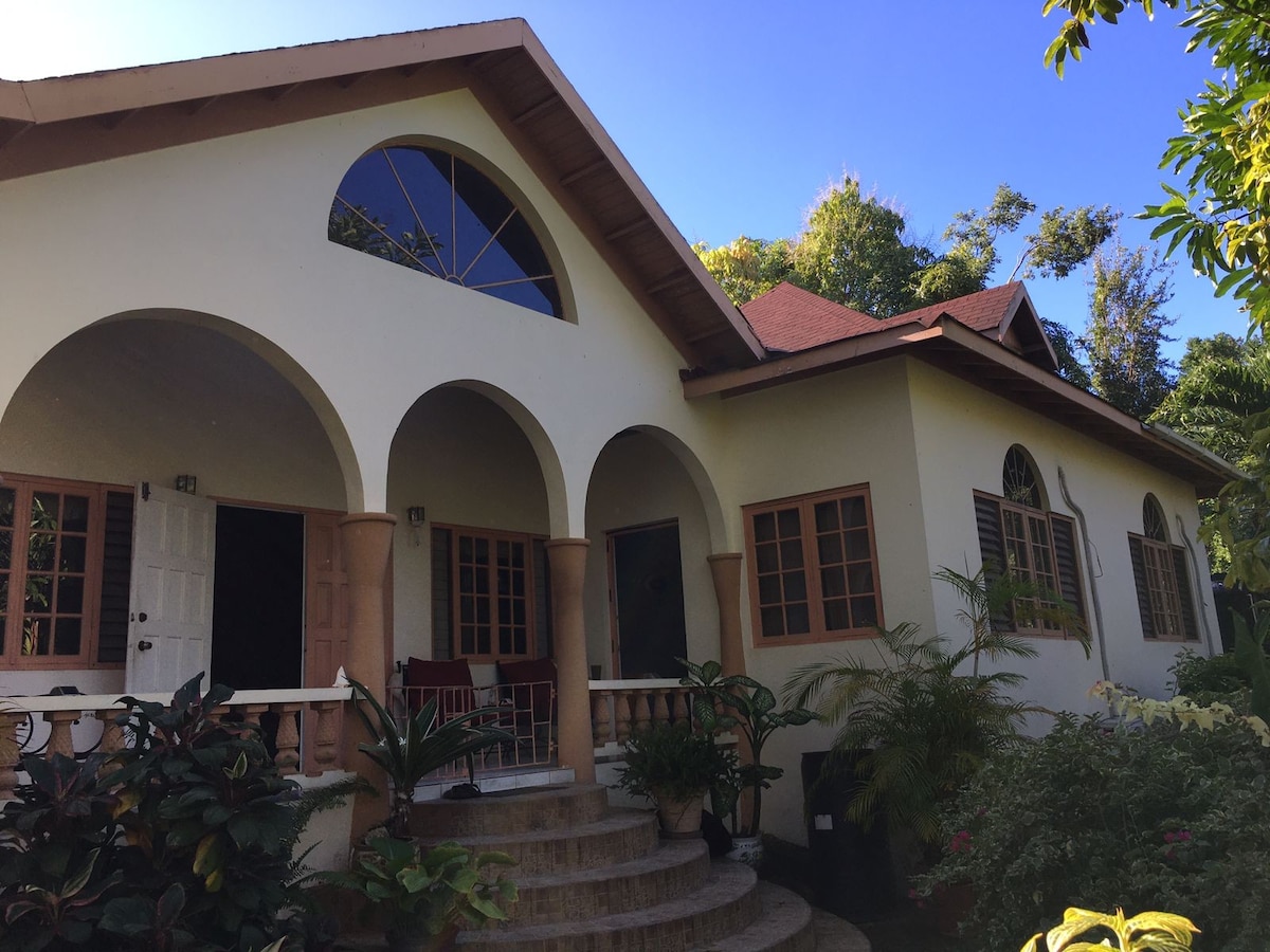 A charming exterior features arched columns and multiple windows, allowing natural light to fill the space. Lush greenery surrounds the entrance, enhancing the tranquil setting. A staircase leads to a welcoming porch, complemented by comfortable seating arrangements.