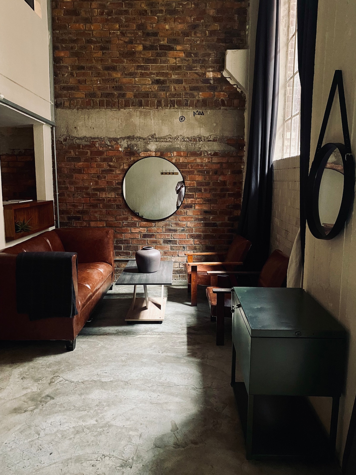 A cozy lounge area features brown leather sofas and wooden accent chairs arranged around a low coffee table. An oval mirror reflects the space, while a grey vase sits on the table. Exposed brick walls and large windows contribute to the loft’s industrial aesthetic.