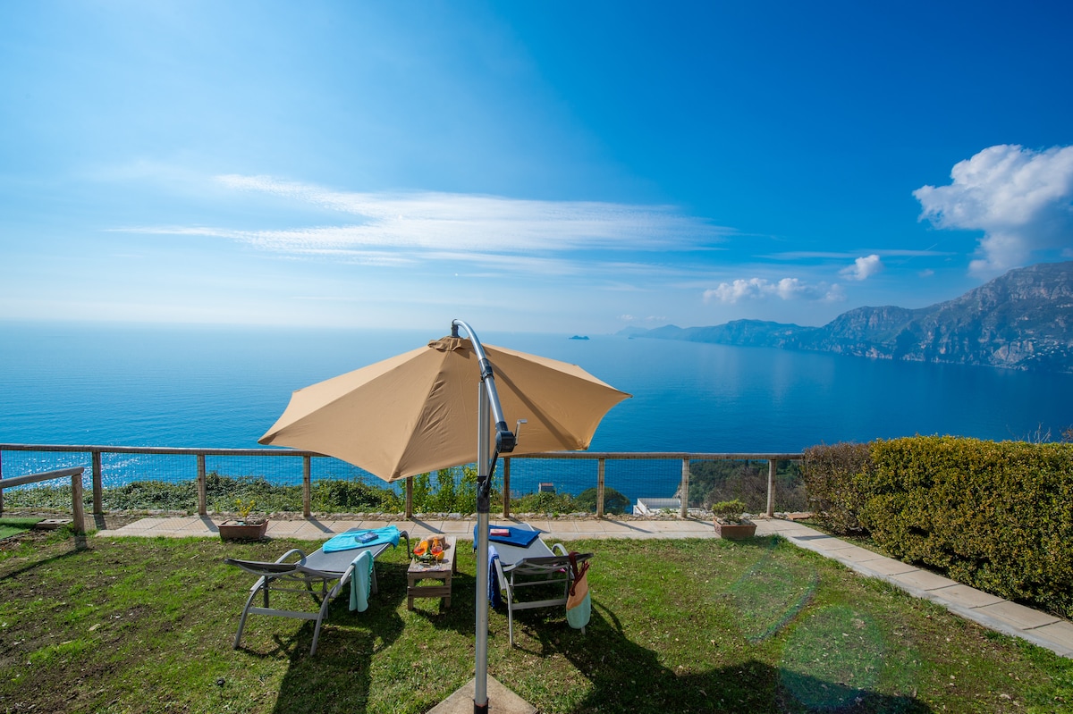 An outdoor solarium area is shown, featuring a large umbrella providing shade over two lounge chairs. The grass is well-maintained, with a view of the expansive sea and distant coastal cliffs under a clear blue sky, conveying a tranquil ambiance.