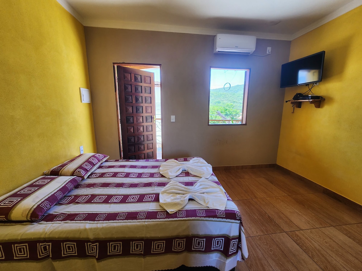 A sunlit bedroom features a spacious bed adorned with patterned bedding. A wooden door opens to a scenic view, while a window allows natural light to enhance the warm yellow walls. A wall-mounted television and air conditioning unit are also visible.