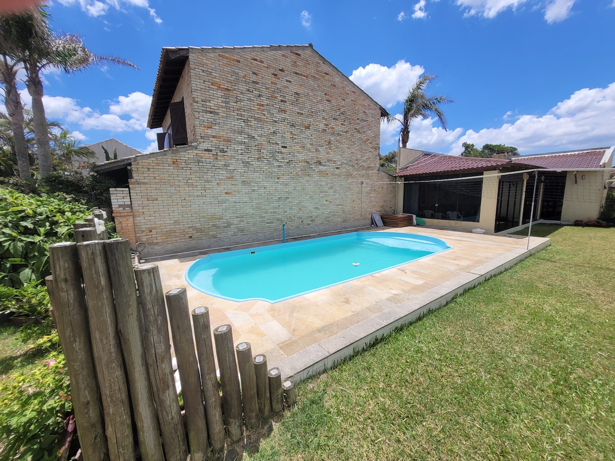 A large swimming pool is surrounded by a spacious patio and well-maintained grass. Lush greenery is visible beyond the pool area, providing a natural setting. A brick wall enhances privacy, and palm trees gently sway in the background under the blue sky.