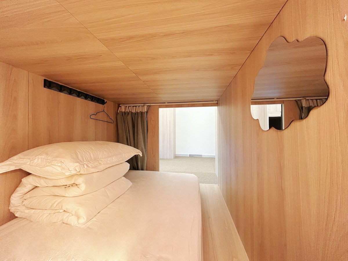 An interior view of a capsule showcases a spacious sleeping area with soft pillows stacked neatly. Wooden paneling creates a warm ambiance, while a small mirror and privacy curtains enhance the space's functionality and comfort.
