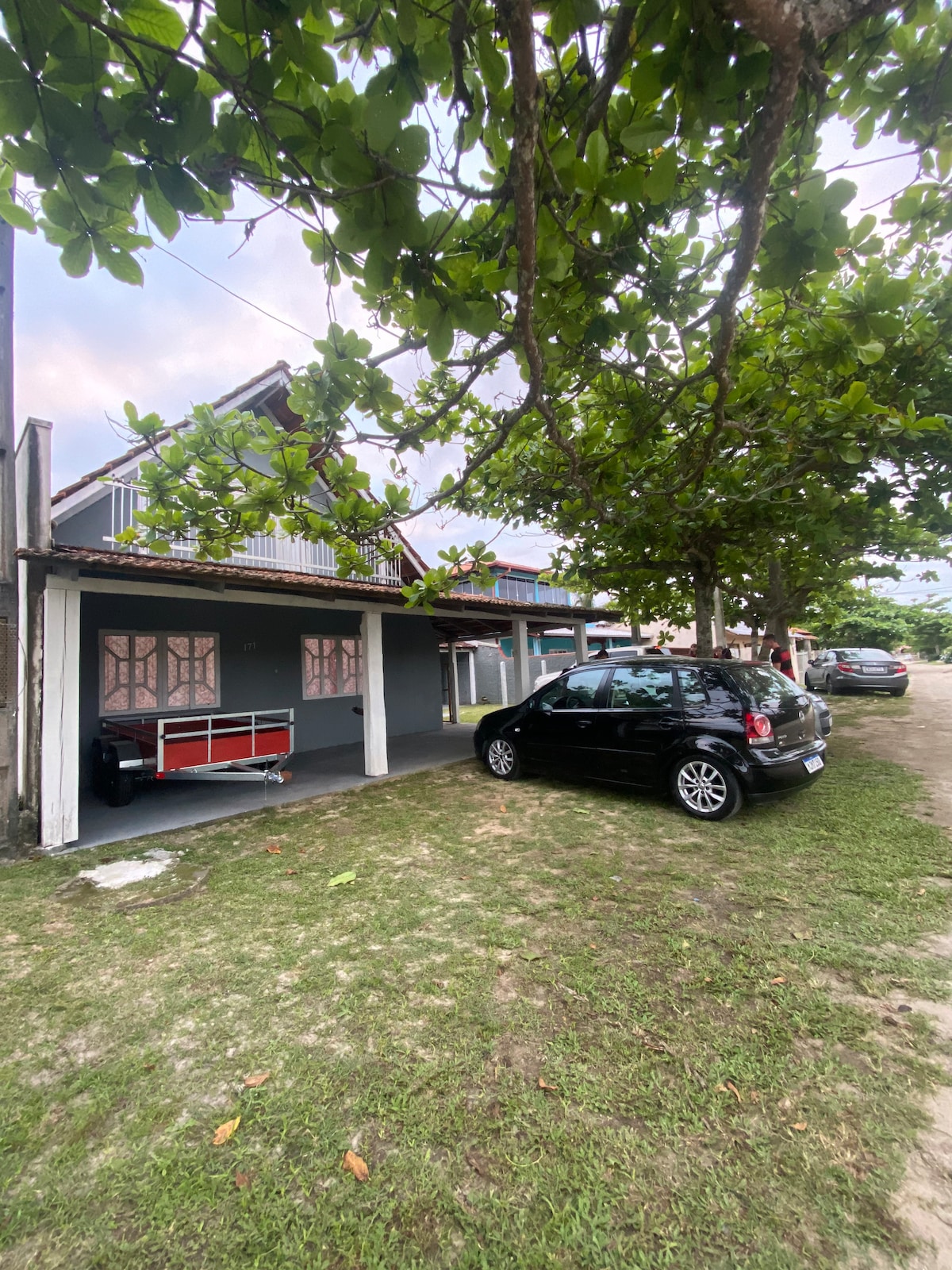 A view of the exterior area showcases a grassy space bordered by trees. A compact car is parked next to a covered outdoor area, which features a storage unit. The building is designed in a simple style, with neighboring structures visible in the background.