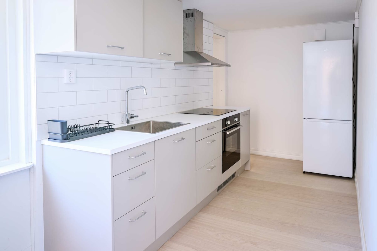 A modern kitchen features sleek cabinetry with a neutral finish. Stainless steel appliances are positioned efficiently, including a refrigerator and an oven. A double sink is integrated into the countertop, and ample counter space is visible, complemented by light-colored flooring that enhances the airy feel.