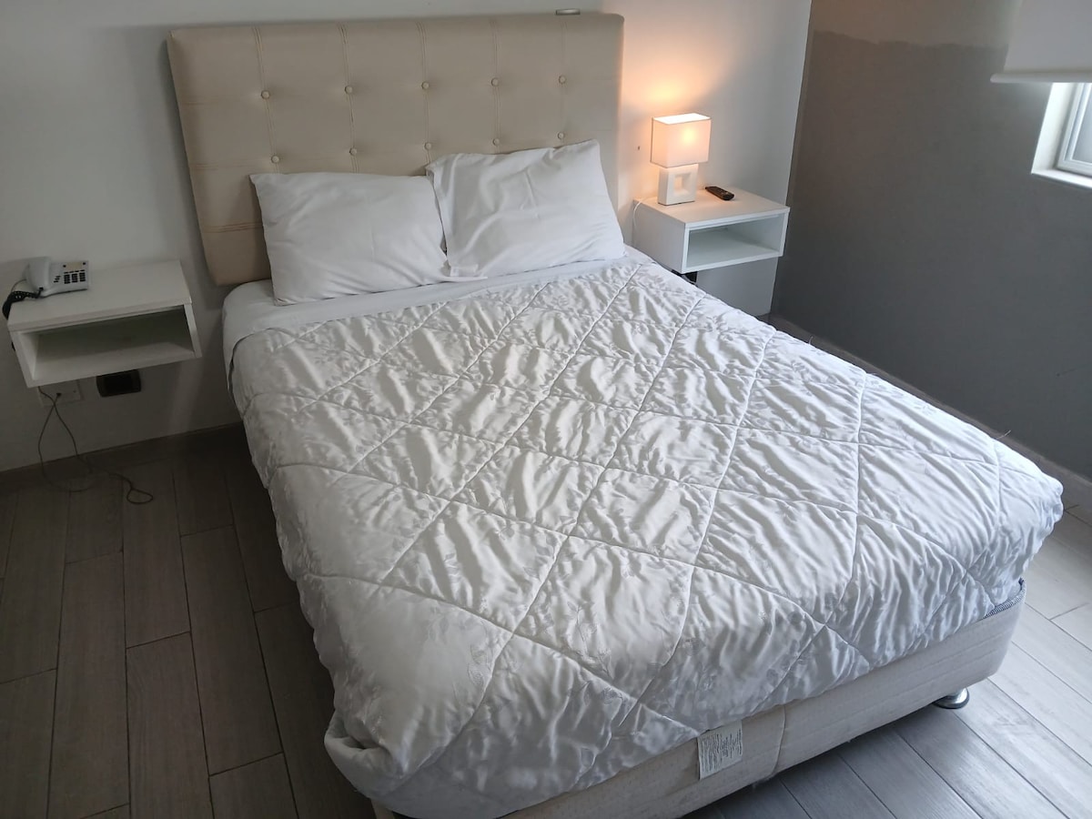 A neatly made bed is centered in the room, featuring a light-colored quilted comforter. Two bedside tables hold simple lamps, providing soft lighting. A window allows natural light to enter, with one wall painted a subtle grey.