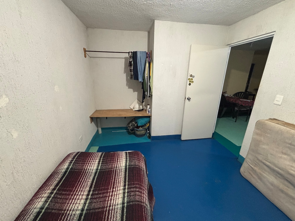 A simple room features a spacious bed covered with a plaid blanket. A wooden shelf holds several articles of clothing, while a basket sits on the floor nearby. A door leads to an adjoining space, and the walls are painted in muted tones.