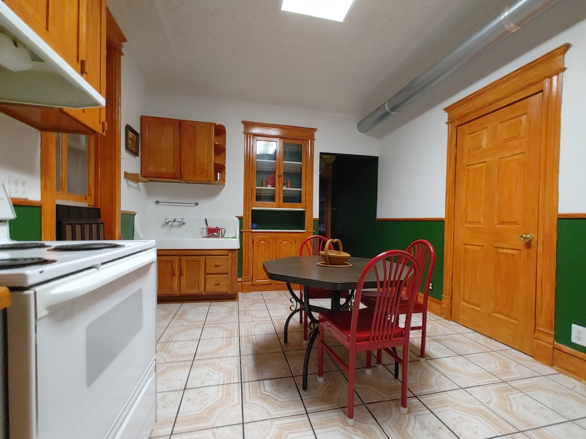 A kitchen area is depicted featuring wooden cabinetry and a white stove. A round table with red chairs occupies the center, complemented by tiled flooring. Light is provided by a ceiling skylight, and green walls add a touch of color to the space.