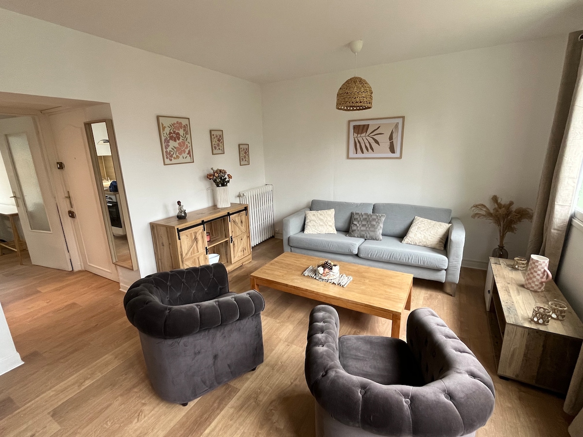 A light-filled living room features a comfortable grey sofa accompanied by two dark armchairs. A wooden coffee table sits in the center, surrounded by decorative accents on side tables. Wall art and flower arrangements add subtle touches to the welcoming atmosphere.