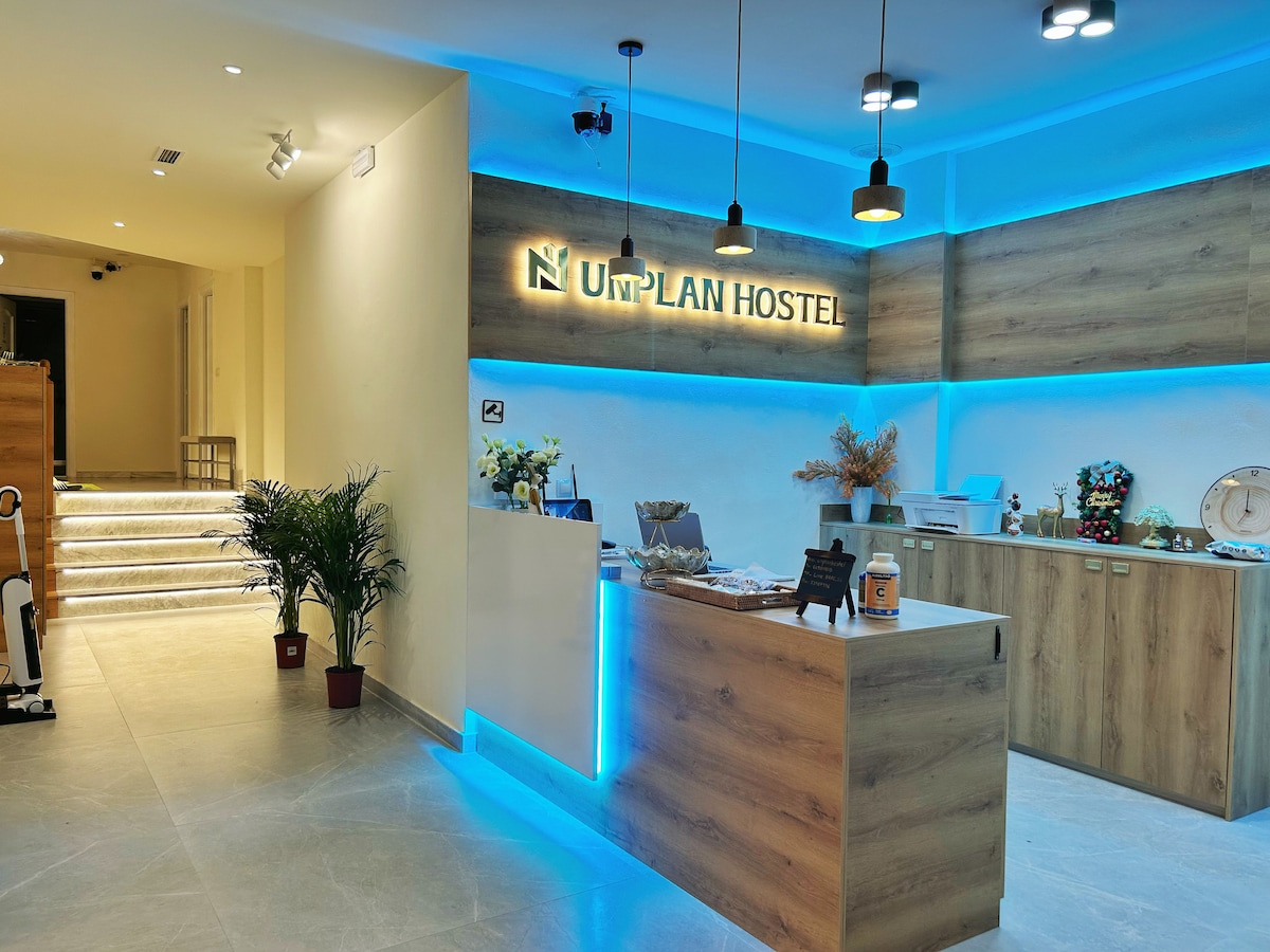 The reception area is adorned with modern lighting that creates a welcoming atmosphere. A wooden front desk is complemented by a blue illuminated panel featuring the hostel's name. Decorative plants and a seating area are visible, enhancing the functionality of the space.