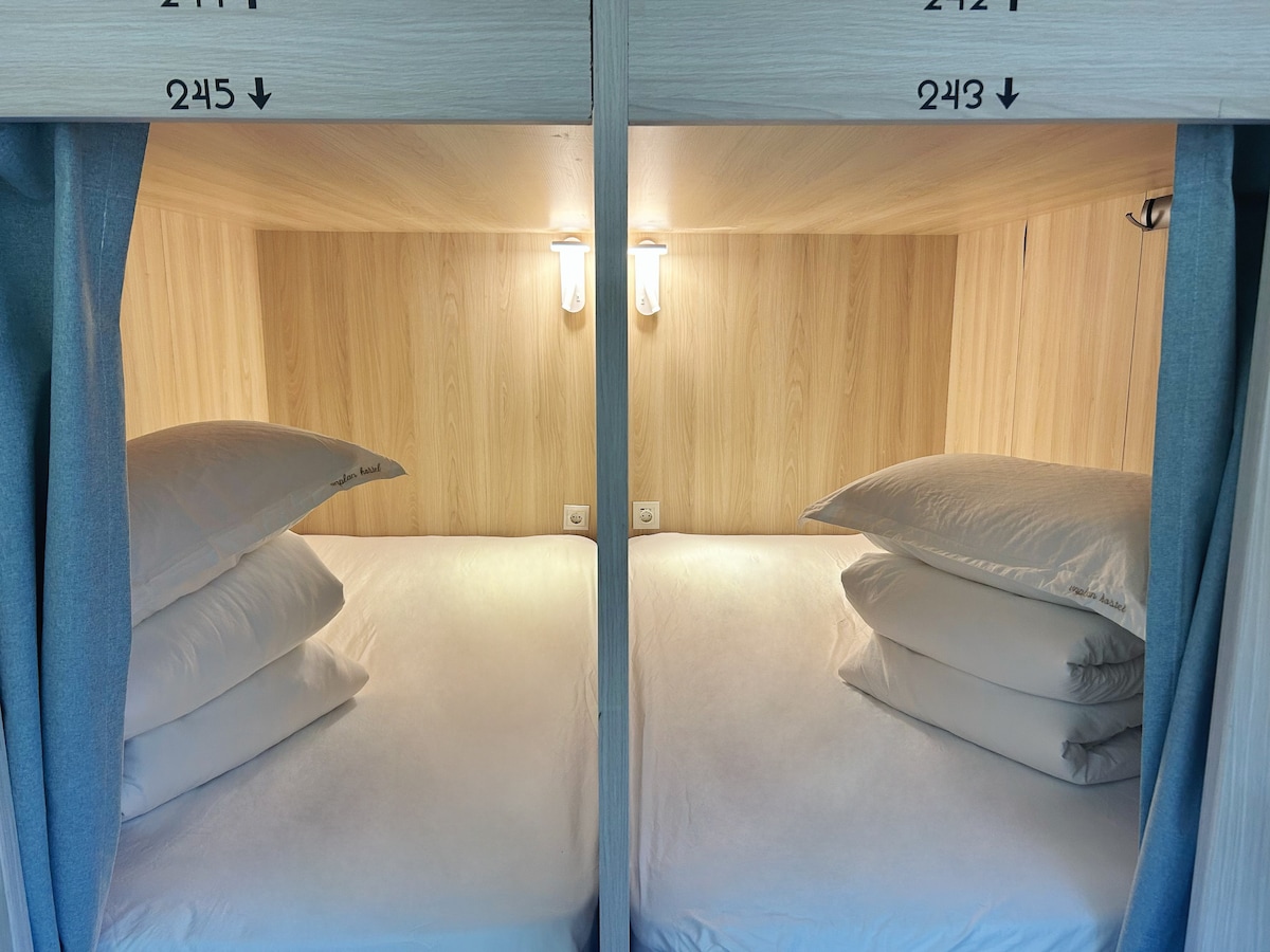 Two capsule beds are arranged side by side, each with a stack of soft pillows. Warm lighting is installed above the beds, and the wooden paneling enhances the serene atmosphere. Privacy curtains are drawn to create an intimate space for each guest.