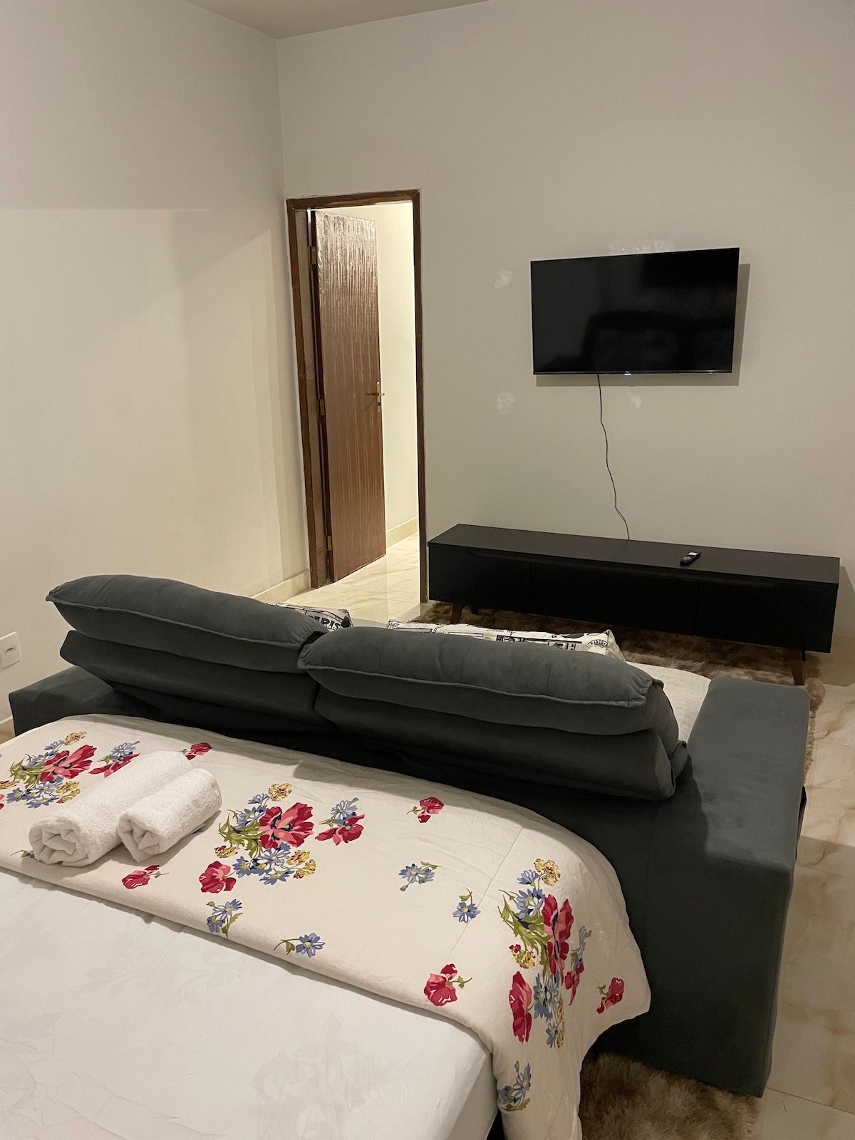 A cozy seating area is furnished with a grey couch and floral throw, positioned in front of a mounted flat-screen TV. A minimalistic black media console rests beneath the TV. A soft rug covers the floor, contributing to a comfortable environment.