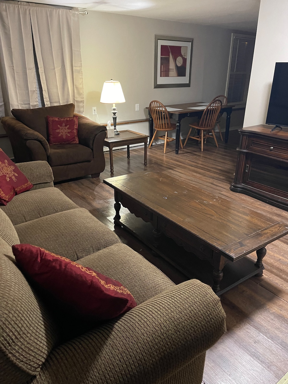 A comfortable living area features a brown couch with decorative cushions and an armchair. A wooden coffee table anchors the space, with a lamp providing soft illumination. A dining table with two chairs is positioned in the background, contributing to the cozy setting.