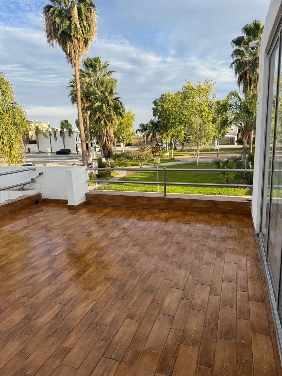 A spacious terrace features wooden flooring and is surrounded by lush greenery and palm trees. Large glass doors provide access to the interior and a clear view of the well-maintained green area outside, creating an open and airy environment.