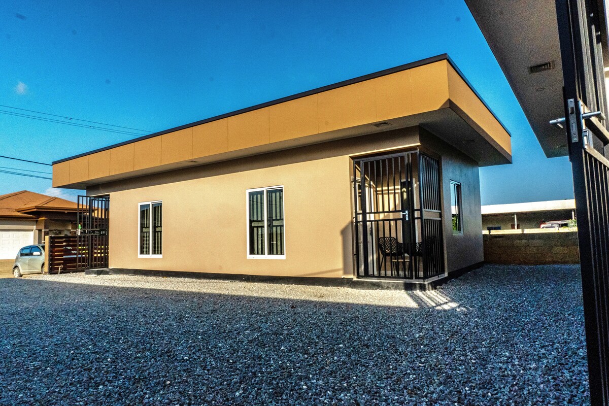 The modern studio apartment is highlighted by a sleek exterior characterized by neutral tones and large windows. A covered porch is visible, featuring outdoor seating. The surrounding area is finished with gravel, providing a clean and accessible environment.