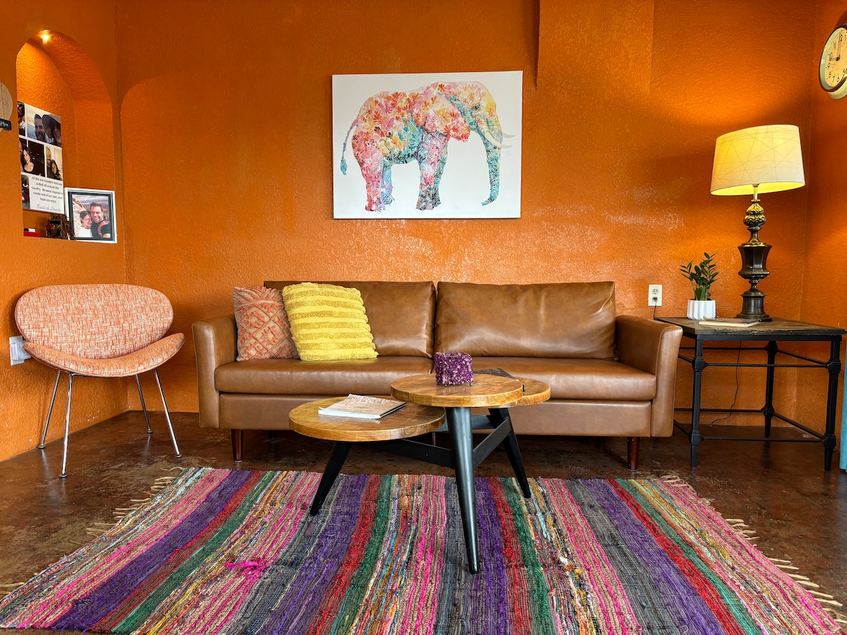 A vibrant living area features an orange wall adorned with an elephant artwork. A brown leather sofa is complemented by a yellow throw blanket. A round wooden coffee table is placed on a colorful striped rug, with a lamp and a vase decorated nearby.