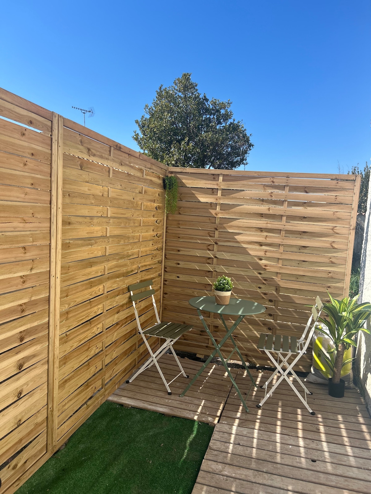 An outdoor seating area features a small round table with two folding chairs, surrounded by wooden privacy slats. A potted plant sits atop the table, and a small patch of artificial grass adds a touch of greenery to the space. A clear blue sky is visible above.